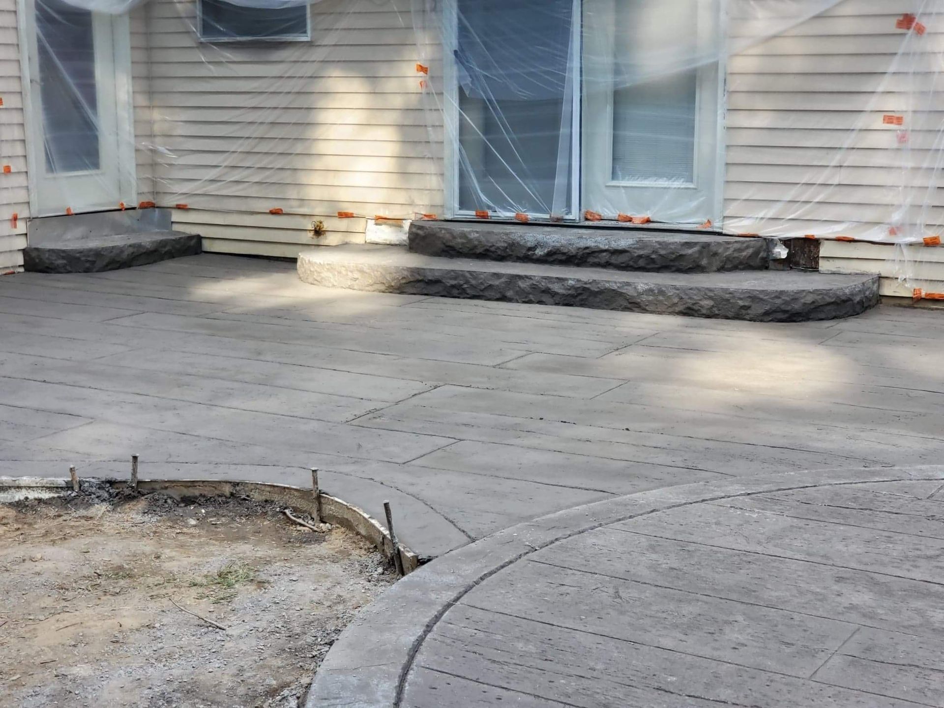 A newly poured gray concrete patio with curved steps leading to two doorways on a house with beige siding.