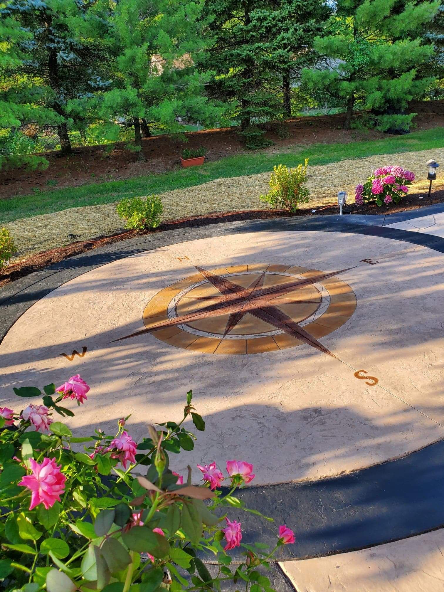 A decorative compass rose embedded in a circular patio, bordered by a dark ring, with pink roses in the foreground.