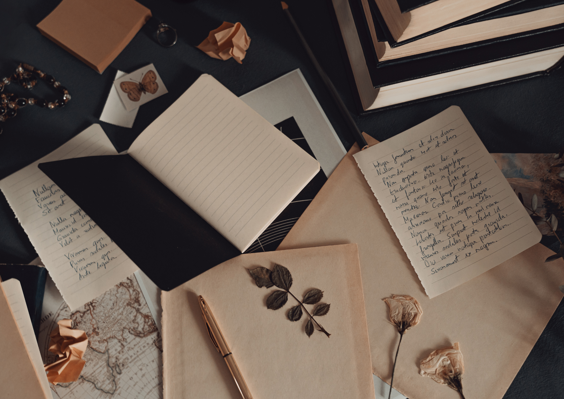 Assortment of notebooks, papers, and dried flowers on a dark surface, suggesting writing and reflection.