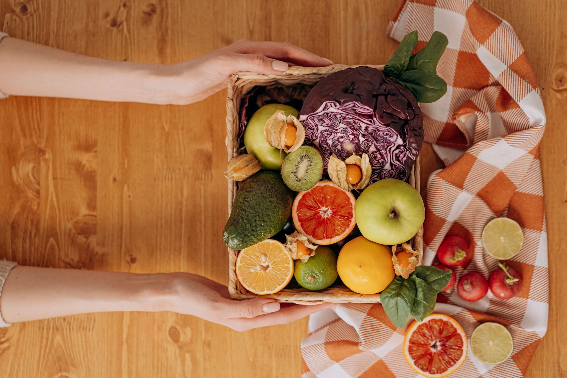 Hands holding a basket filled with colorful fruits and vegetables on a wooden surface next to a patterned cloth.