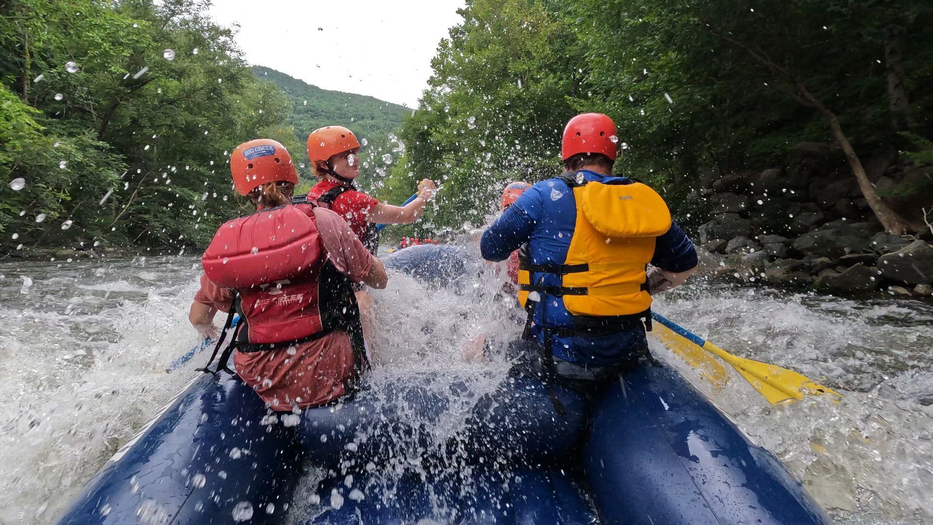 Three people in life vests and helmets white-water raft down a river, surrounded by splashing water and lush trees.