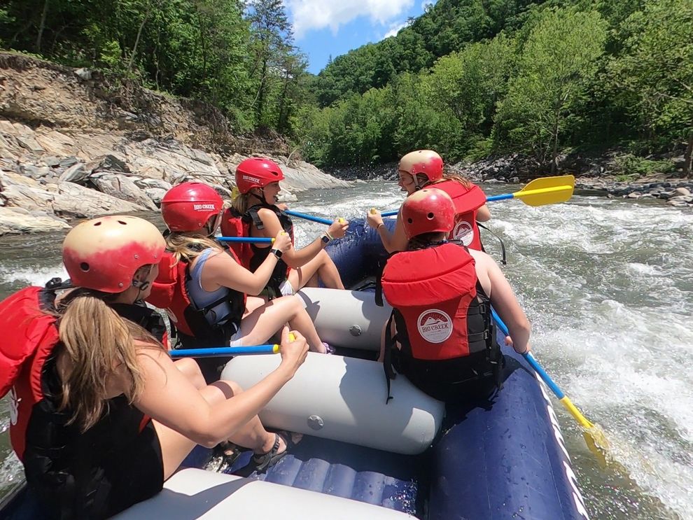 Rafting group in red helmets paddles through whitewater on a river surrounded by rocky, wooded banks
