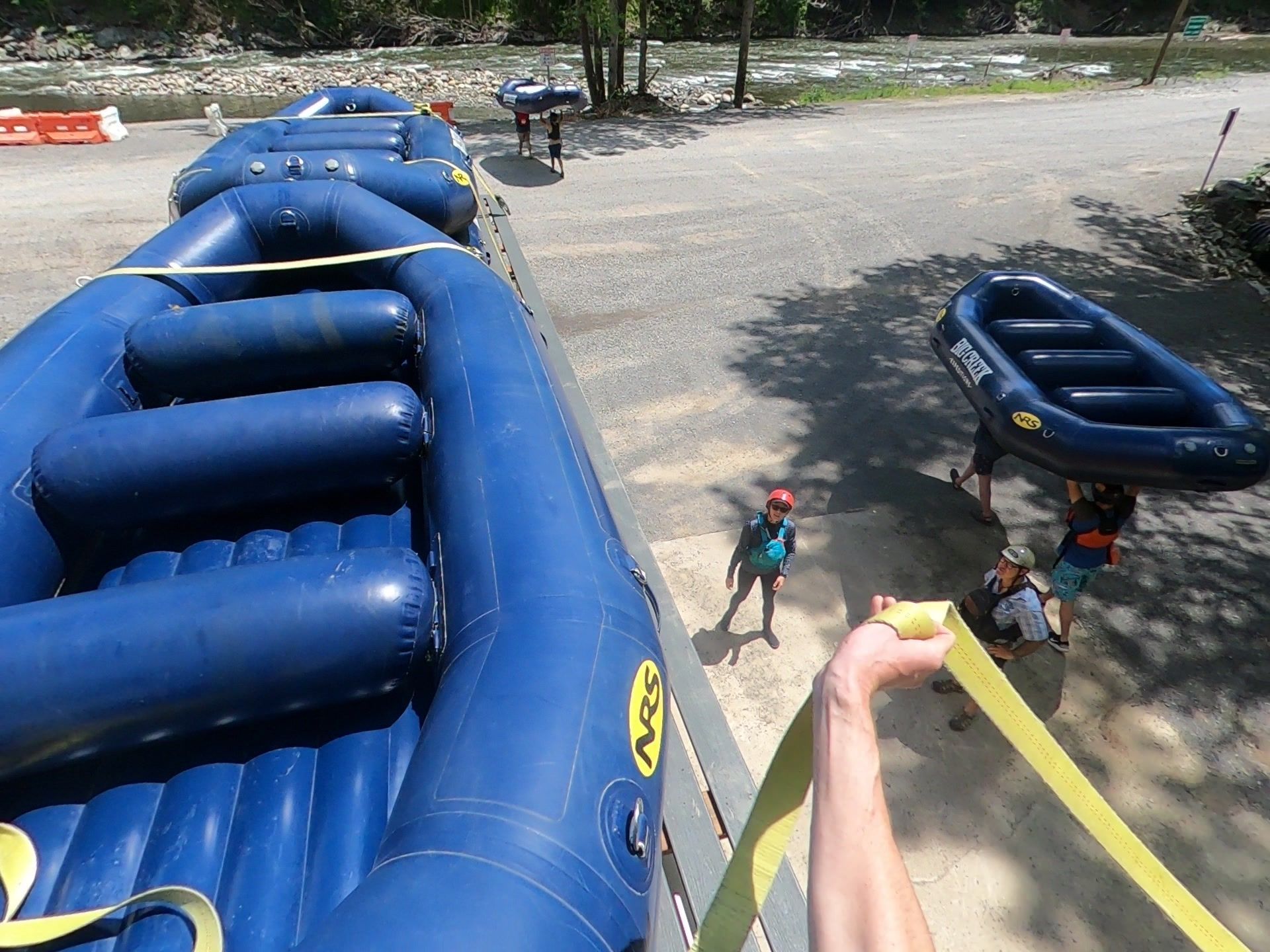 A first-person view of a person securing a large blue inflatable raft with yellow straps near a riverbank.