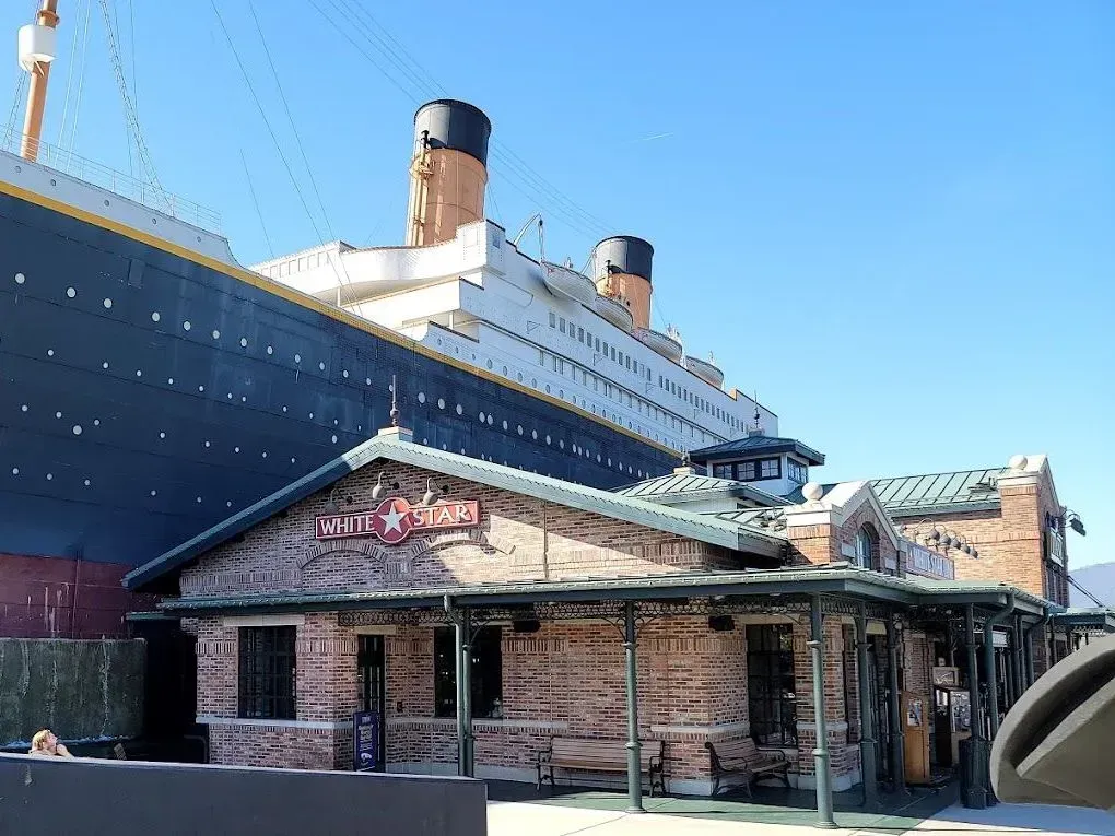 Historic brick building beside the Titanic in bright daylight, with the ship’s hull and smokestacks towering above.