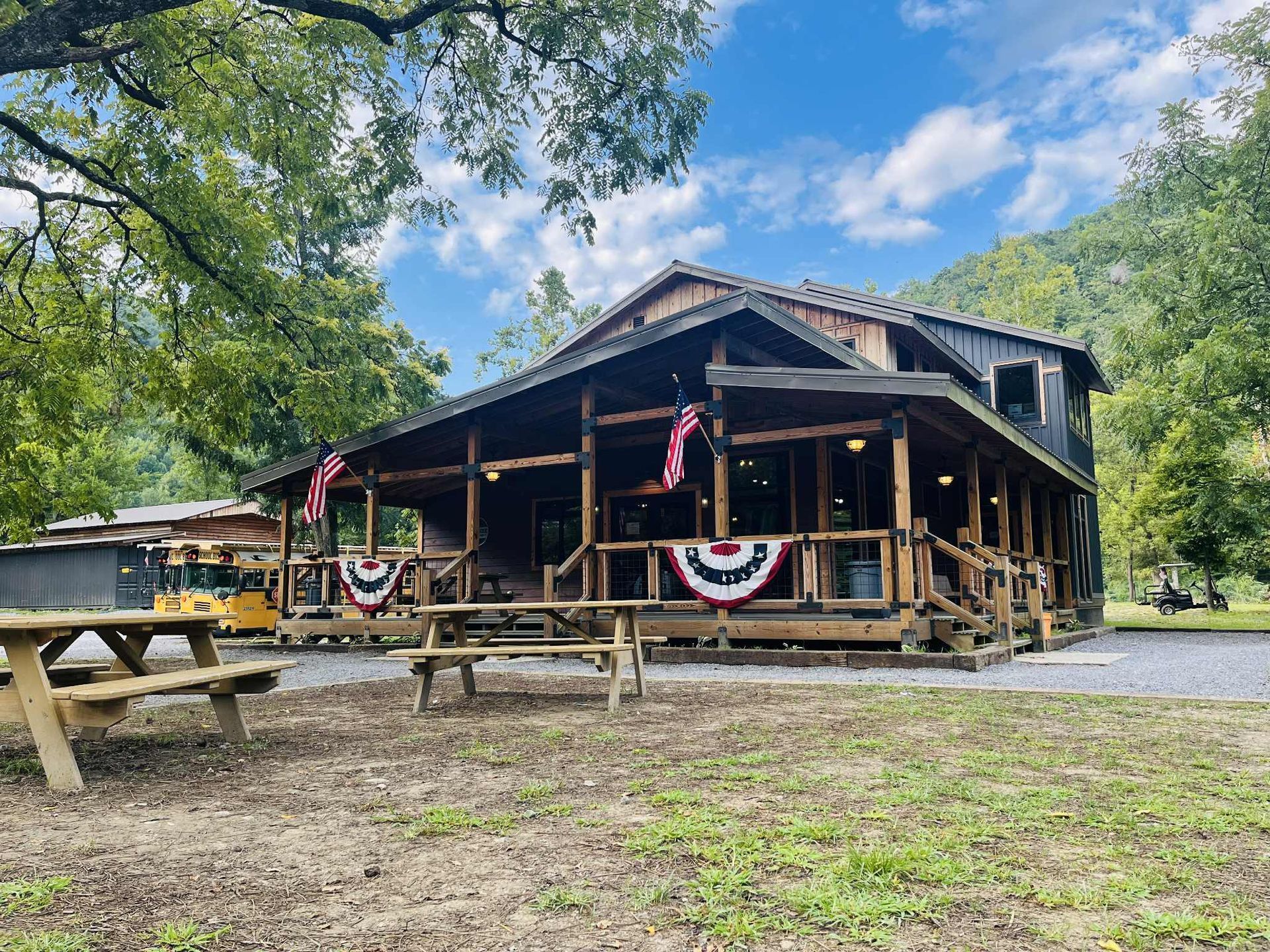 A rustic wooden lodge with a large wrap-around porch decorated with patriotic bunting, set in a forested, hilly area.