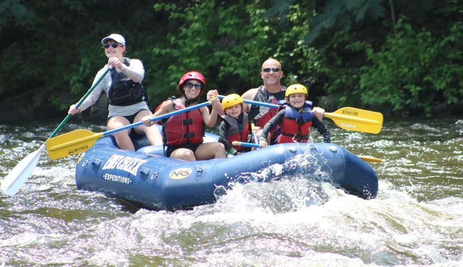 Four people whitewater rafting in a blue raft, paddles raised, splashing through rapids near a forested riverbank
