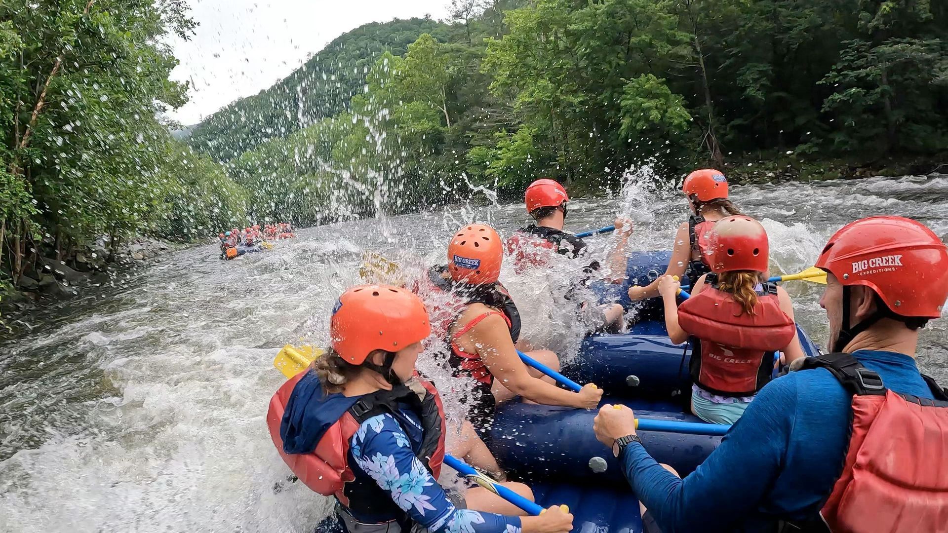 A group of people wearing life vests and helmets white-water rafting down a river with splashing water.