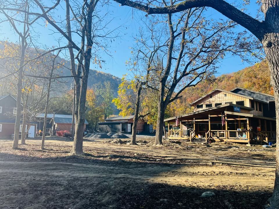 A dirt clearing in a wooded area with trees surrounding a large wooden house and several outbuildings under a blue sky.