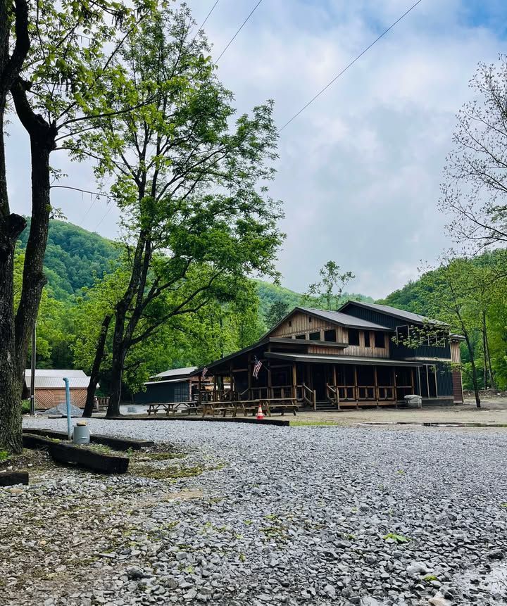 Wooden building with a large porch, set against a backdrop of green mountains and trees under a cloudy sky.