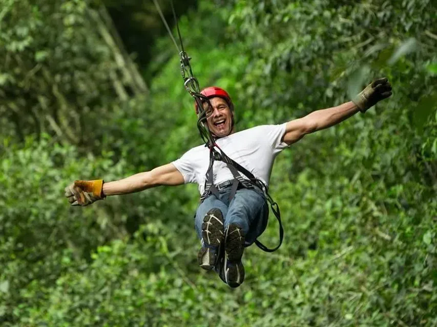 Person ziplining with arms outstretched, wearing a red helmet and white shirt over a green forest.