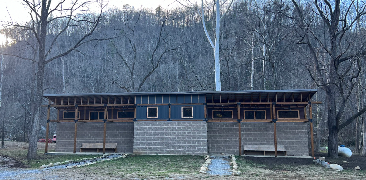 A one-story building with brick walls, a wooden frame porch, and rectangular windows set against a forest background.
