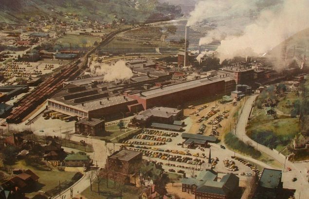 Aerial view of a large, industrial factory complex with smokestacks, train tracks, and a parking lot.