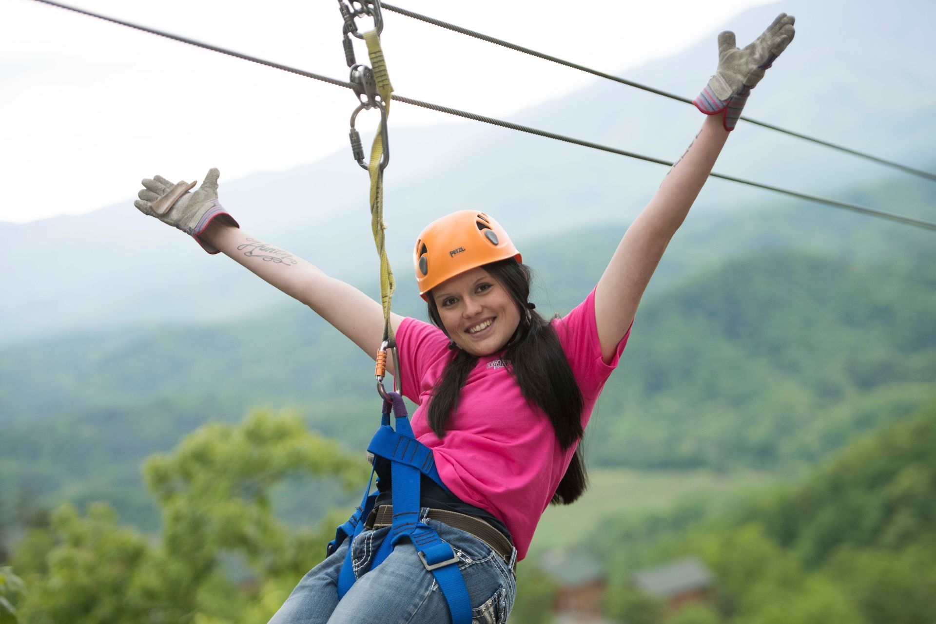 Woman zip-lining with arms raised, wearing an orange helmet and pink shirt over a lush green valley.