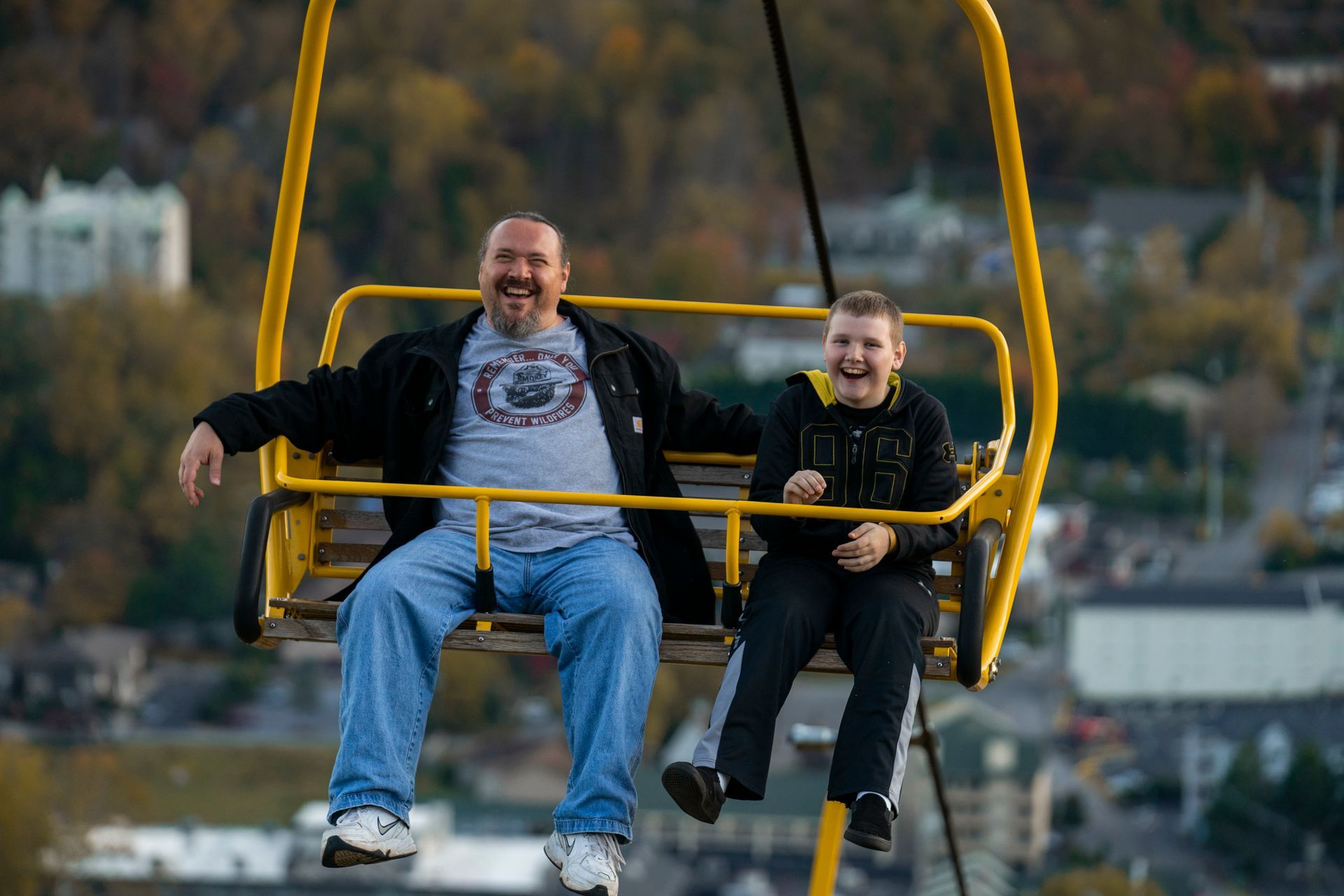 Two people smiling on a yellow ski lift over a blurred town background