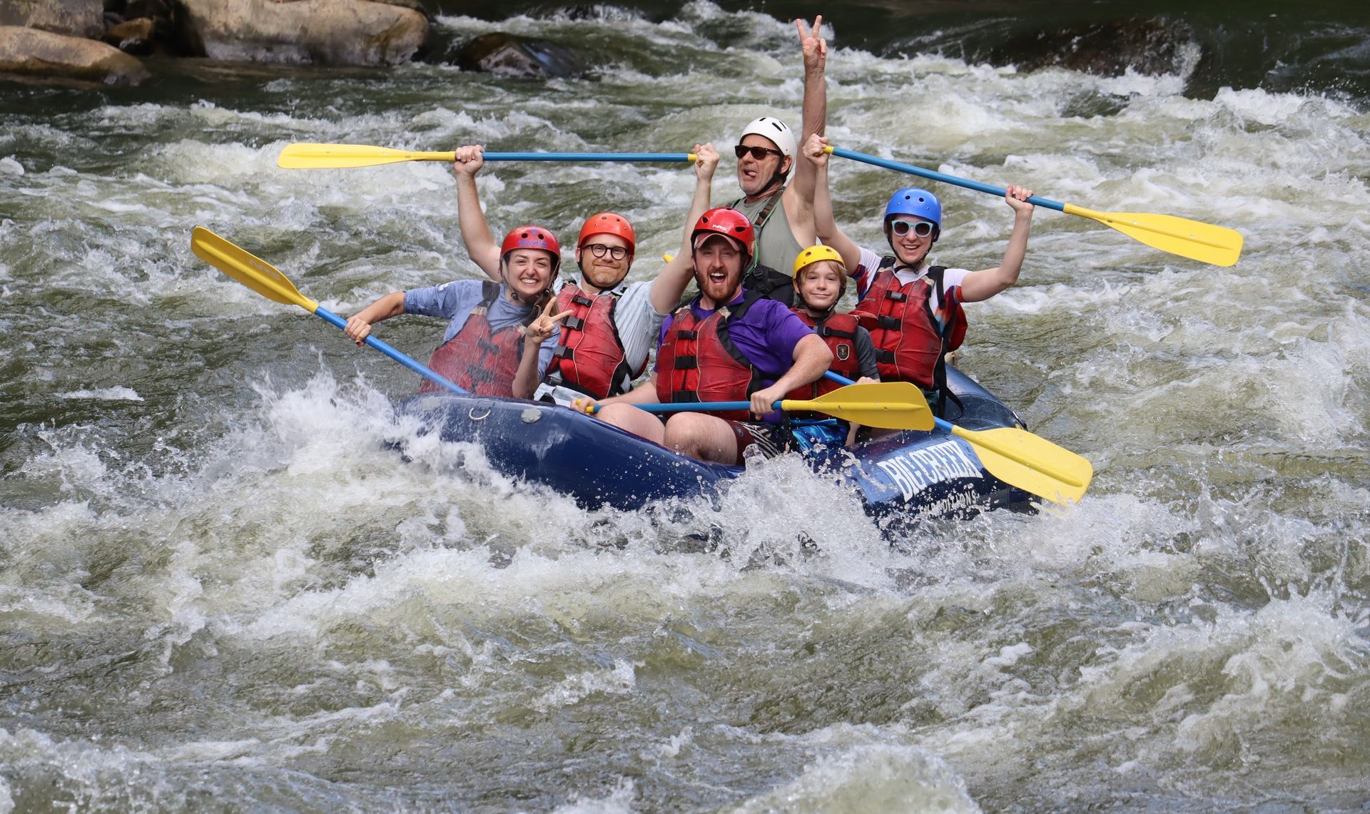 Team of rafters in red helmets paddling through whitewater rapids on a blue inflatable raft