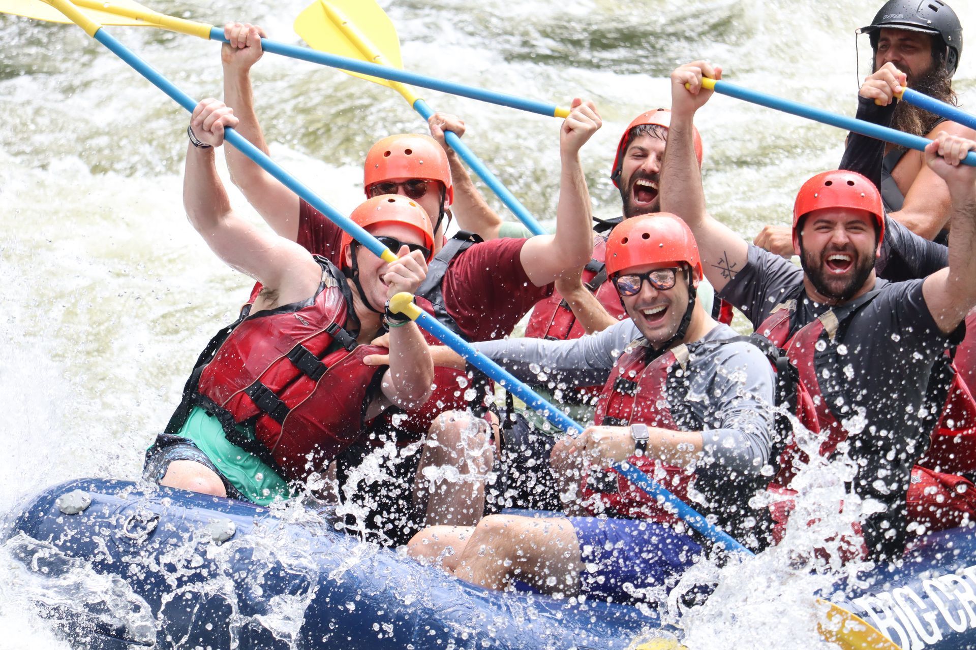 Group whitewater rafters in orange helmets and red vests paddling through splashing rapids