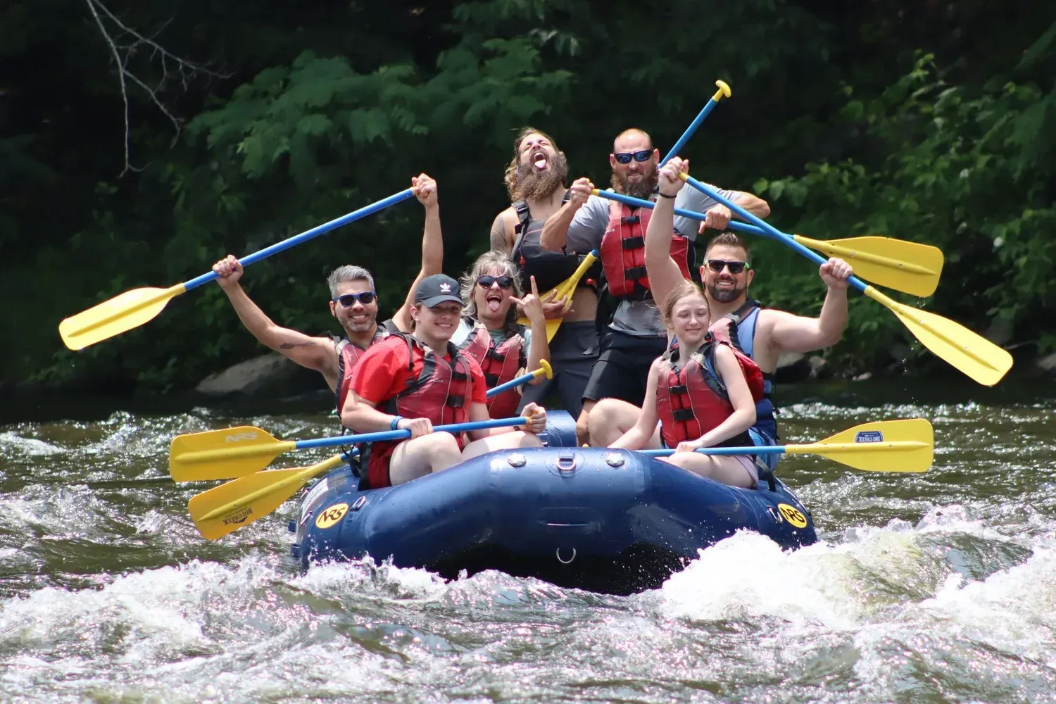 Group whitewater rafting in a blue raft, paddles raised, splashing through rapids