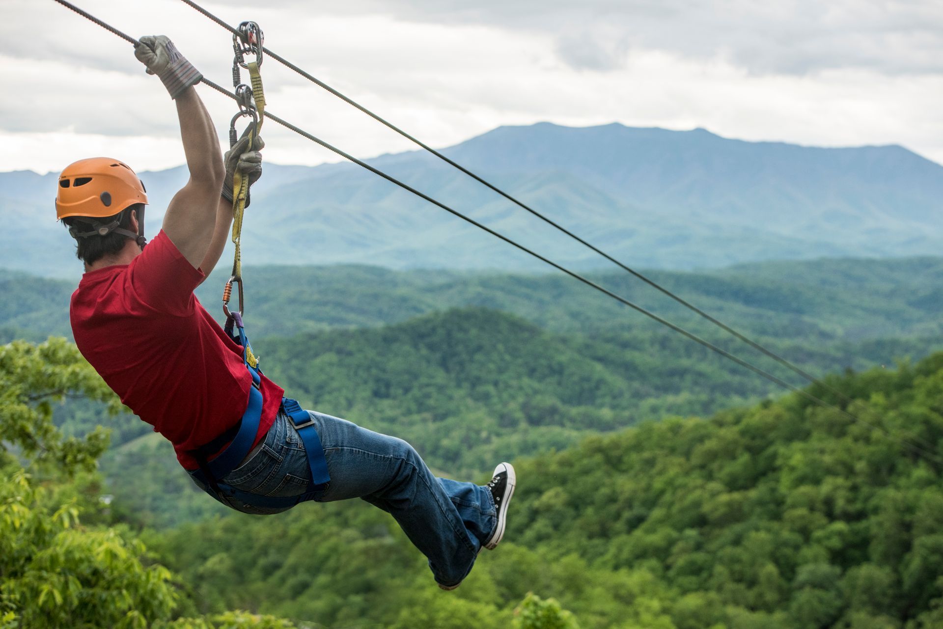 Zipline rider in orange helmet and red shirt glides over a lush green forest with mountains in the background