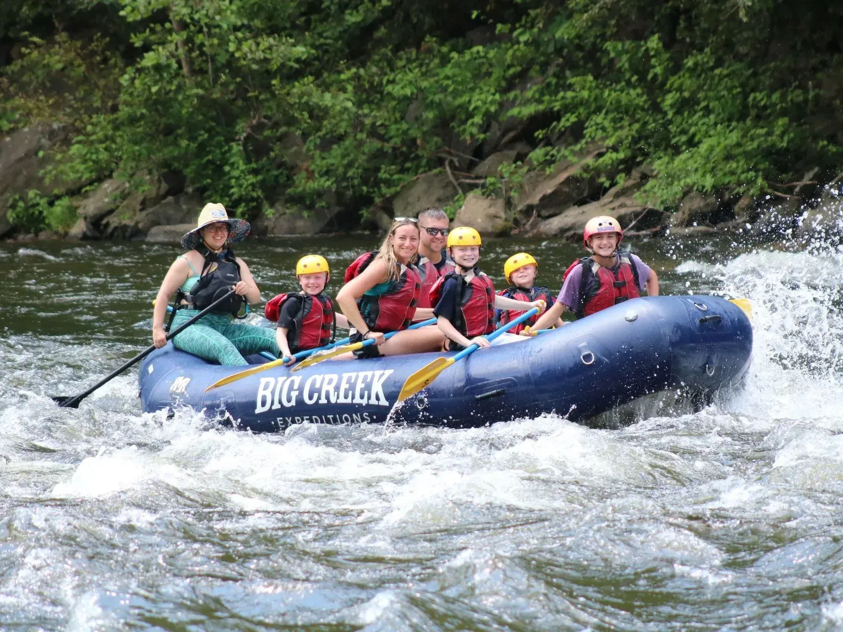 Rafting group in blue raft splashing through whitewater on a river, with green forested banks.
