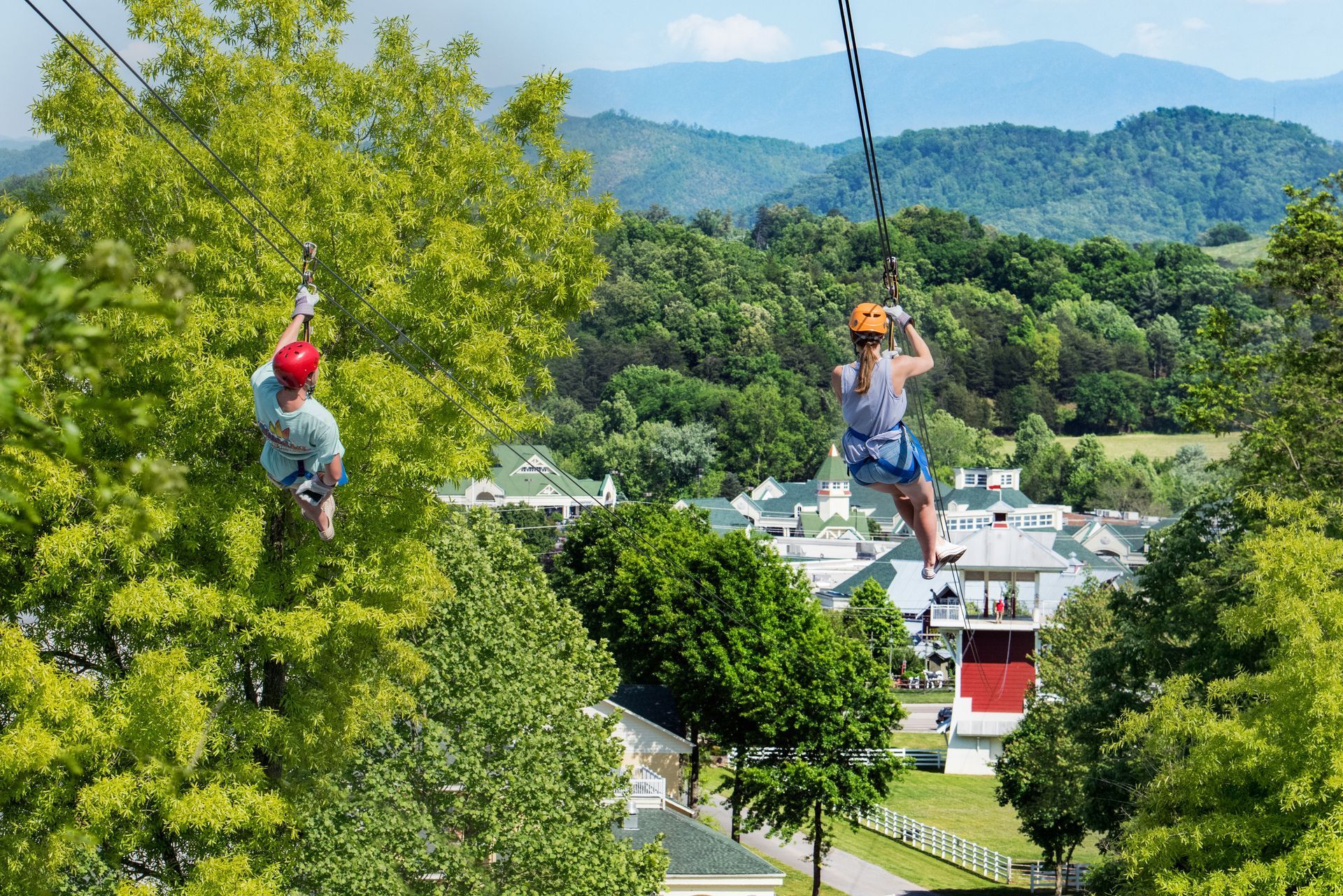 Two people zip-lining over a lush, green valley with buildings and mountains in the distance.