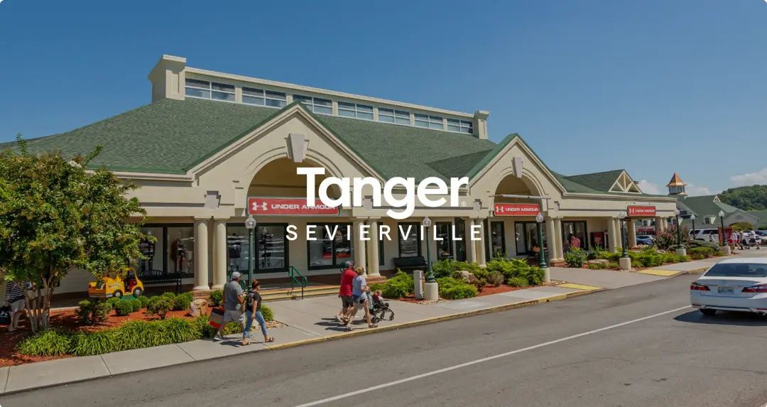 Tanger Outlets storefront with green roof and landscaping on a sunny day, cars parked along the curb