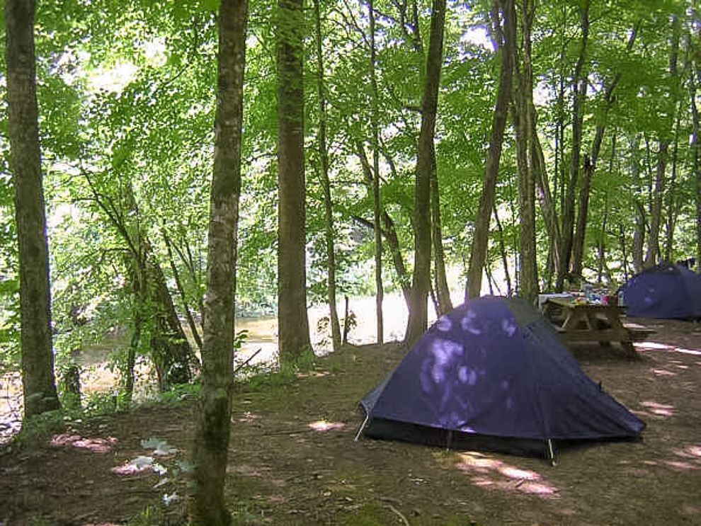 Two dark blue dome tents set up on a dirt clearing in a lush, wooded forest near a body of water.