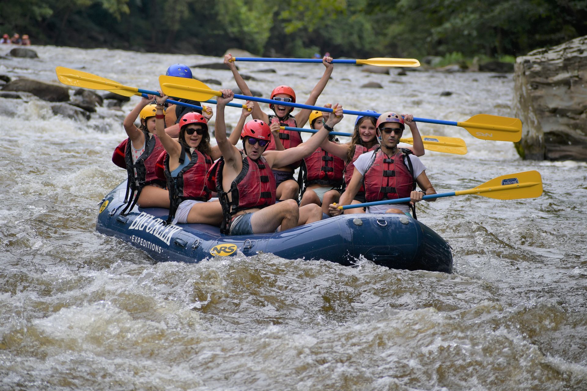 Whitewater raft full of paddlers wearing red life jackets and helmets racing through rapids