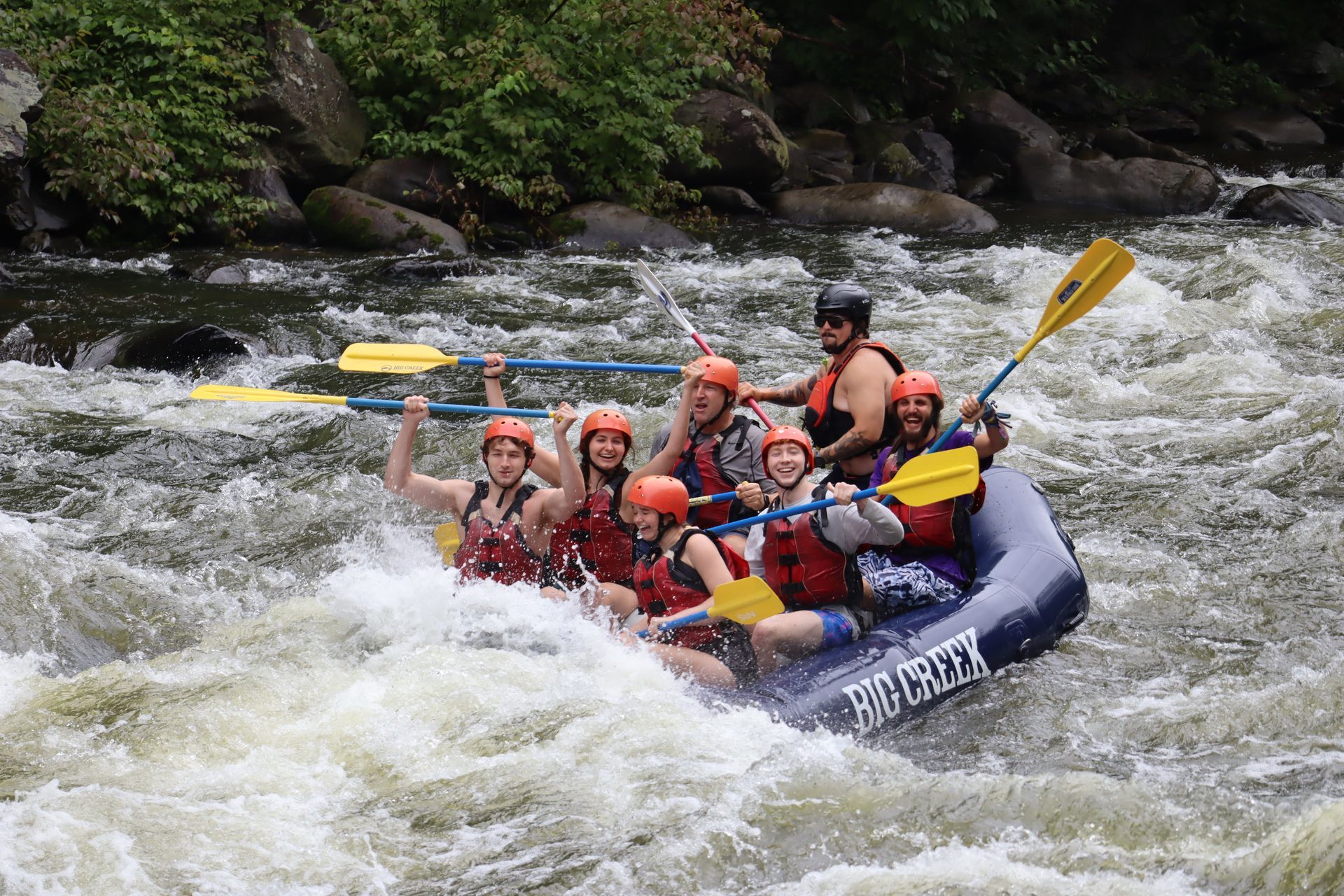 Whitewater rafters in red helmets paddling a blue raft through rough rapids near a rocky forested riverbank