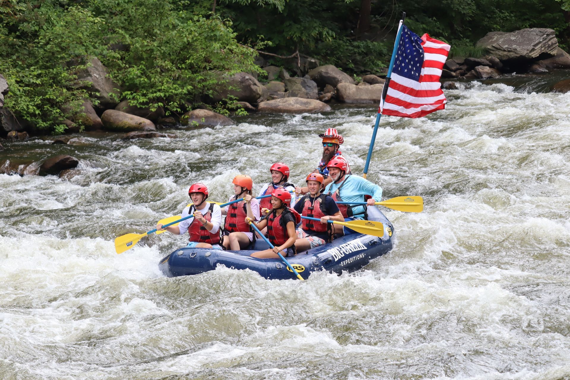 Whitewater rafting group in a blue raft, paddling through rapids with an American flag on the back