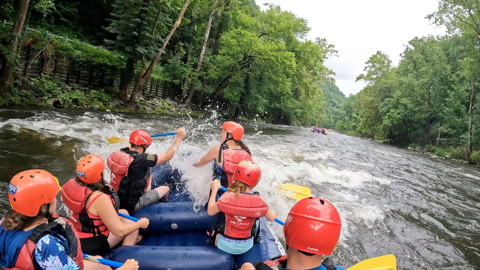 People in red helmets and life vests rafting down a forested river, paddling through splashing water.