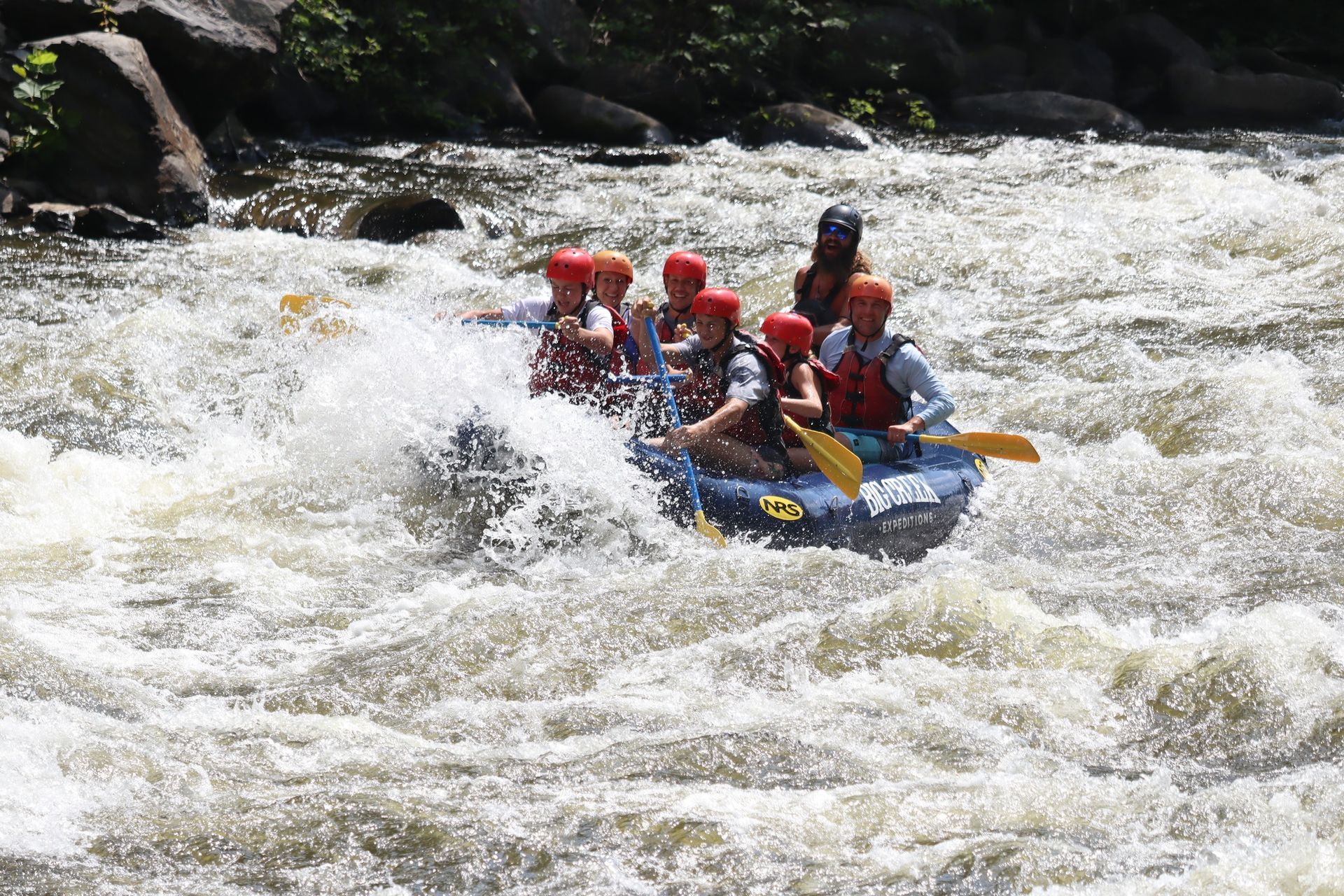 Group whitewater rafting in a blue raft, paddling through rough rapids surrounded by rocky riverbanks