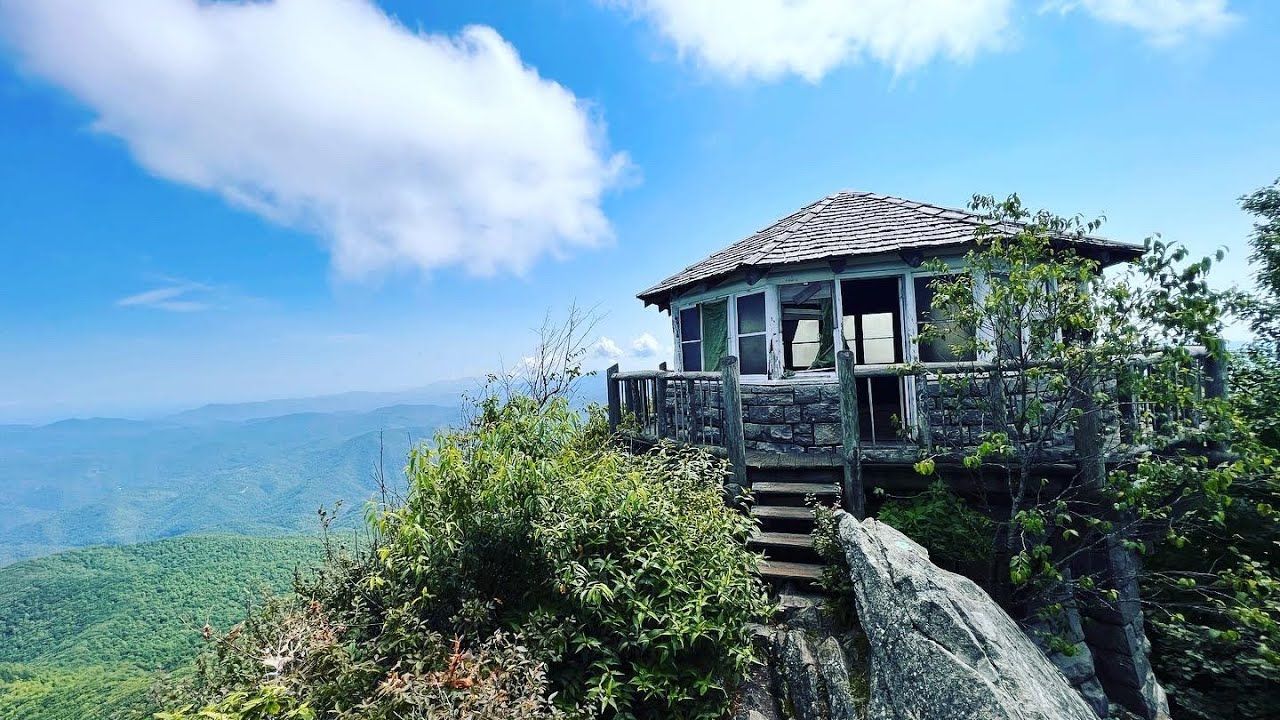 A historic wooden fire lookout tower perched on a rocky mountain summit overlooking lush green valleys under a blue sky.