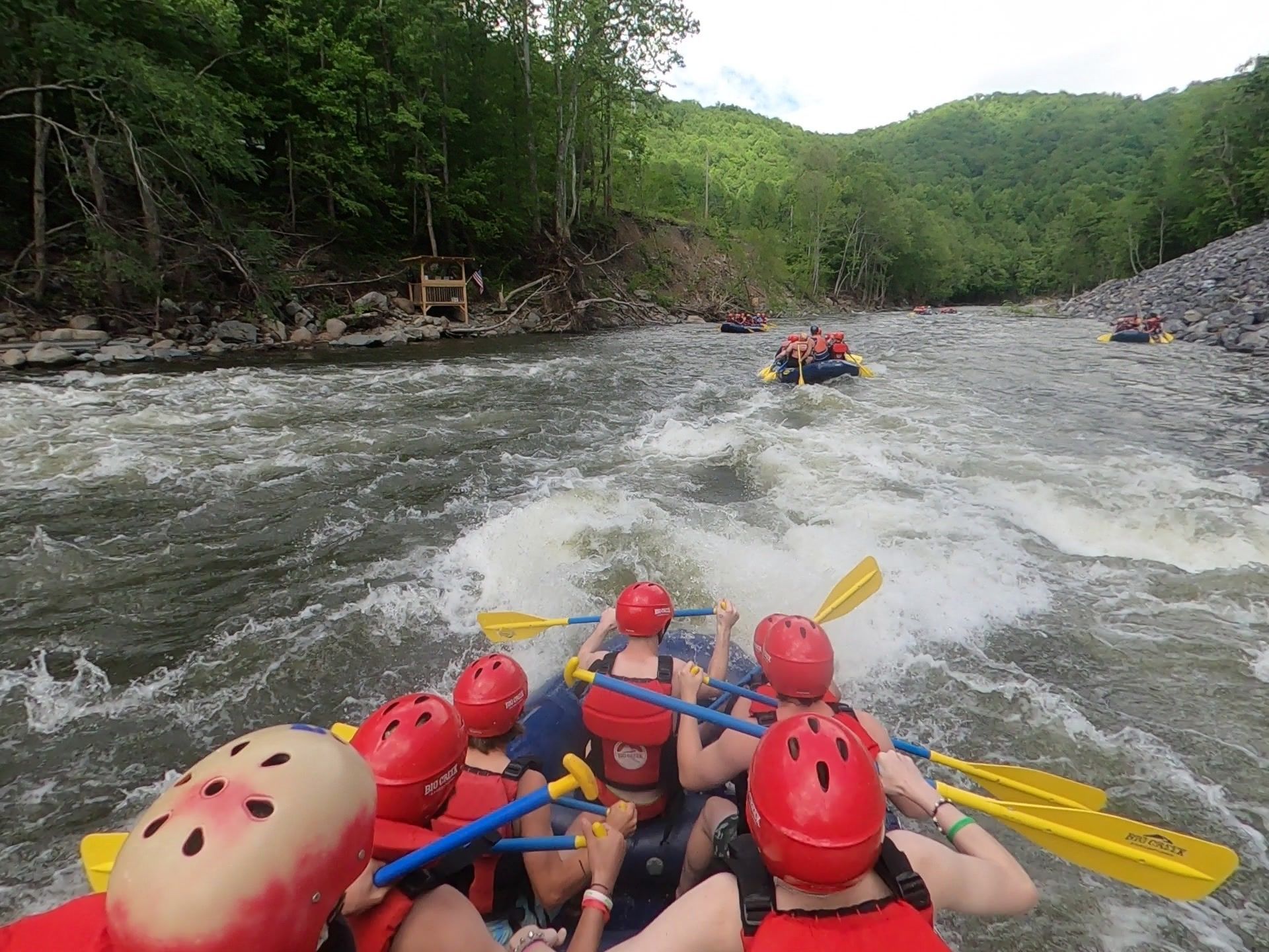 Rafting group in red helmets paddles through rough river rapids, surrounded by green forested hills.