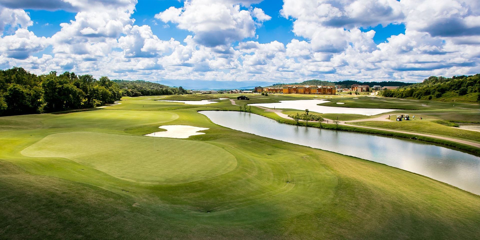 Golf course with green fairways, sand bunkers, and a winding water hazard under a partly cloudy sky
