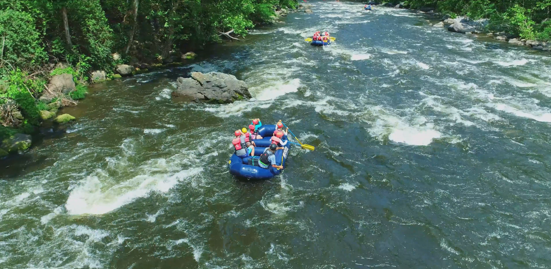 A group of people in a blue raft navigate a river with gentle rapids, surrounded by a lush green forest.