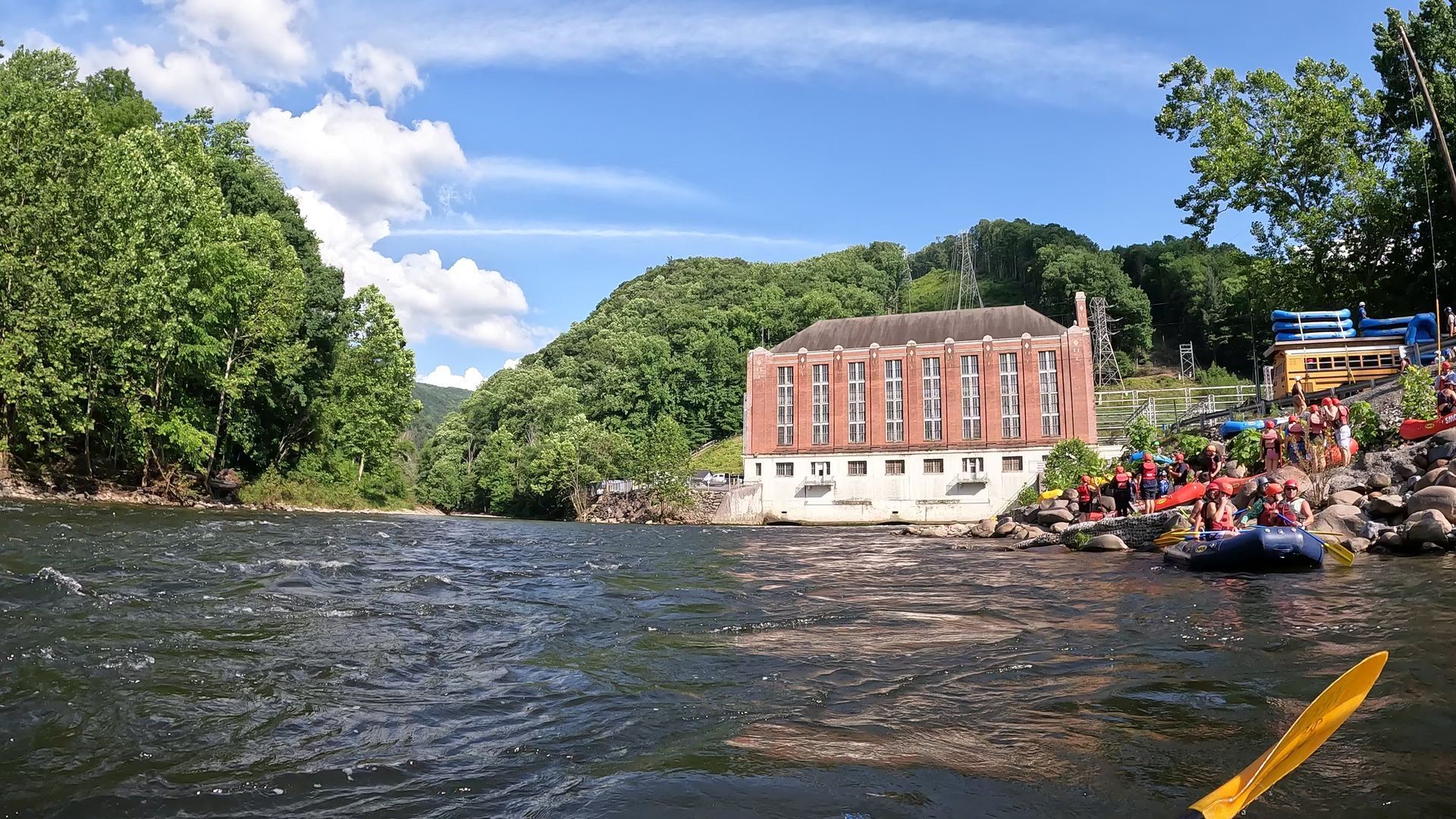A red brick building sits by a river with people in rafts near the shore under a blue sky with lush green hills.