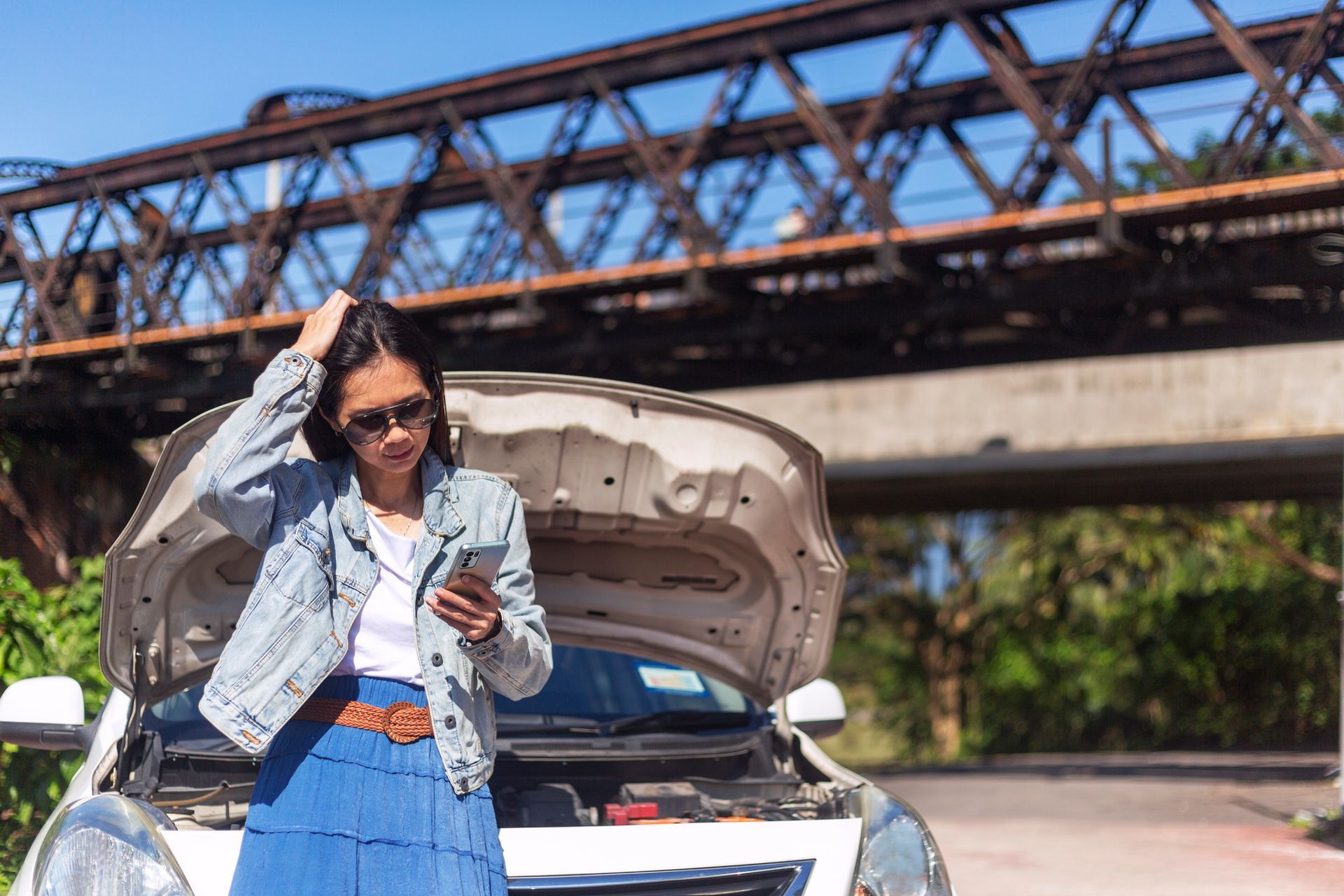 A woman is standing next to a broken down car with the hood up and looking at her phone.
