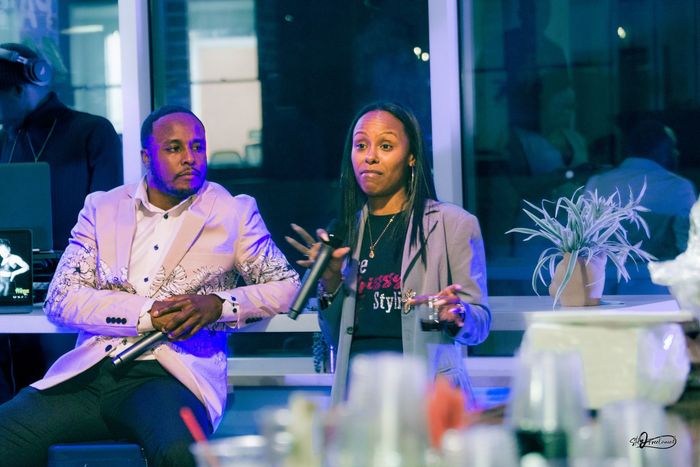 Chris rivers and people speaking at an event with microphones, seated near a window and plant, another person is DJing.
