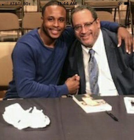 Chris Rivers and a man smiling, arm in arm, at a table with a book. One wears a blue shirt, the other a suit.