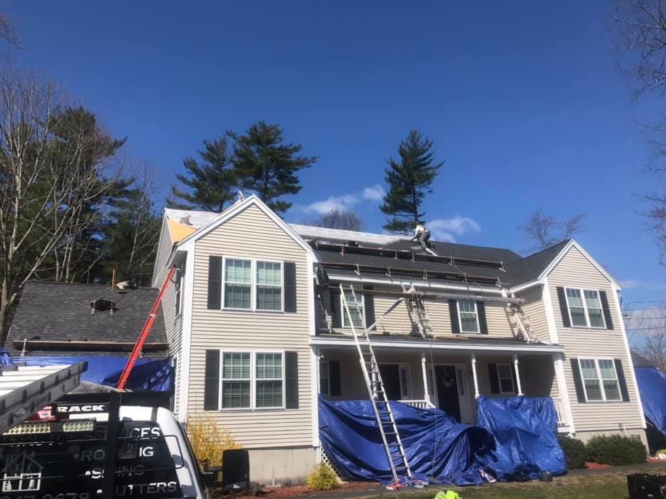 Roof replacement in progress on a two-story house, blue tarps, ladders, and workers on the roof.