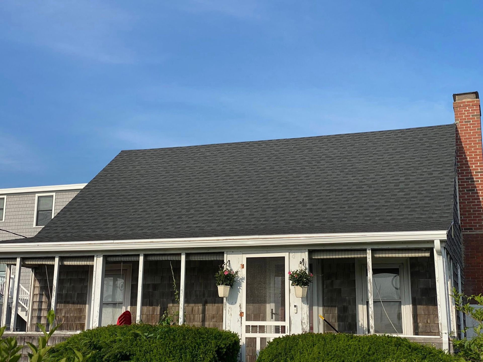 Screened porch of a cottage with dark shingle roof, red brick chimney, and blue sky.