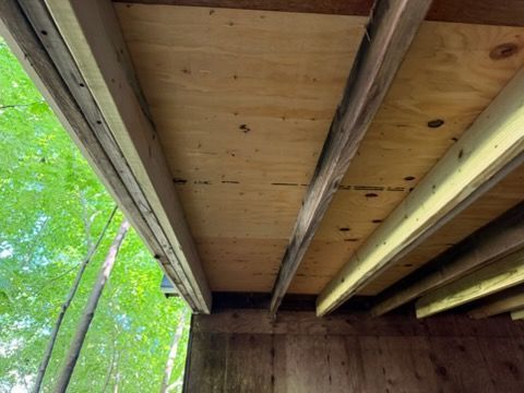 Underneath a wooden deck with beams and plywood, viewed upward toward green trees beyond