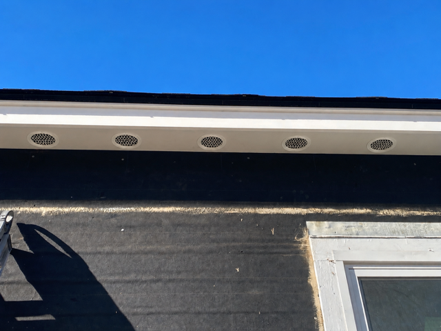 Close-up of a house roof edge with soffit vents against a blue sky, above dark siding and a window