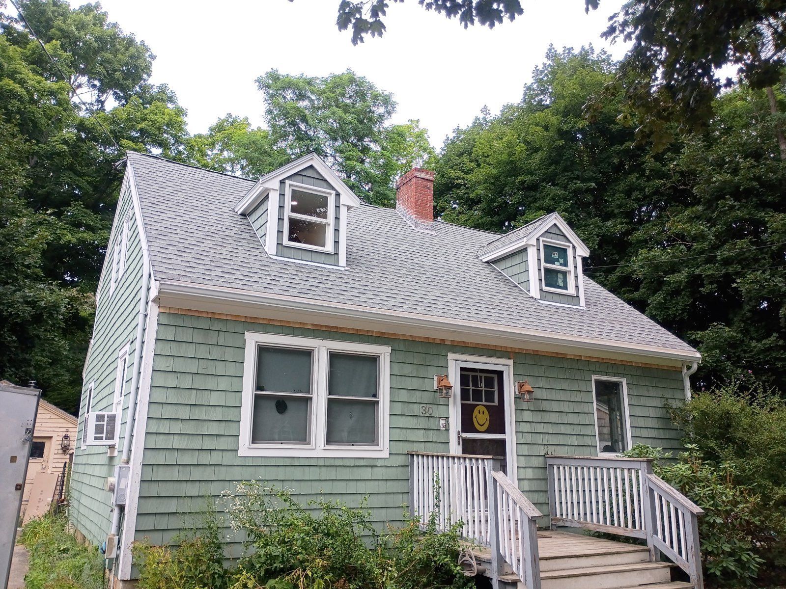 Small light-green house with white trim, front steps, dormer windows, and a chimney surrounded by trees