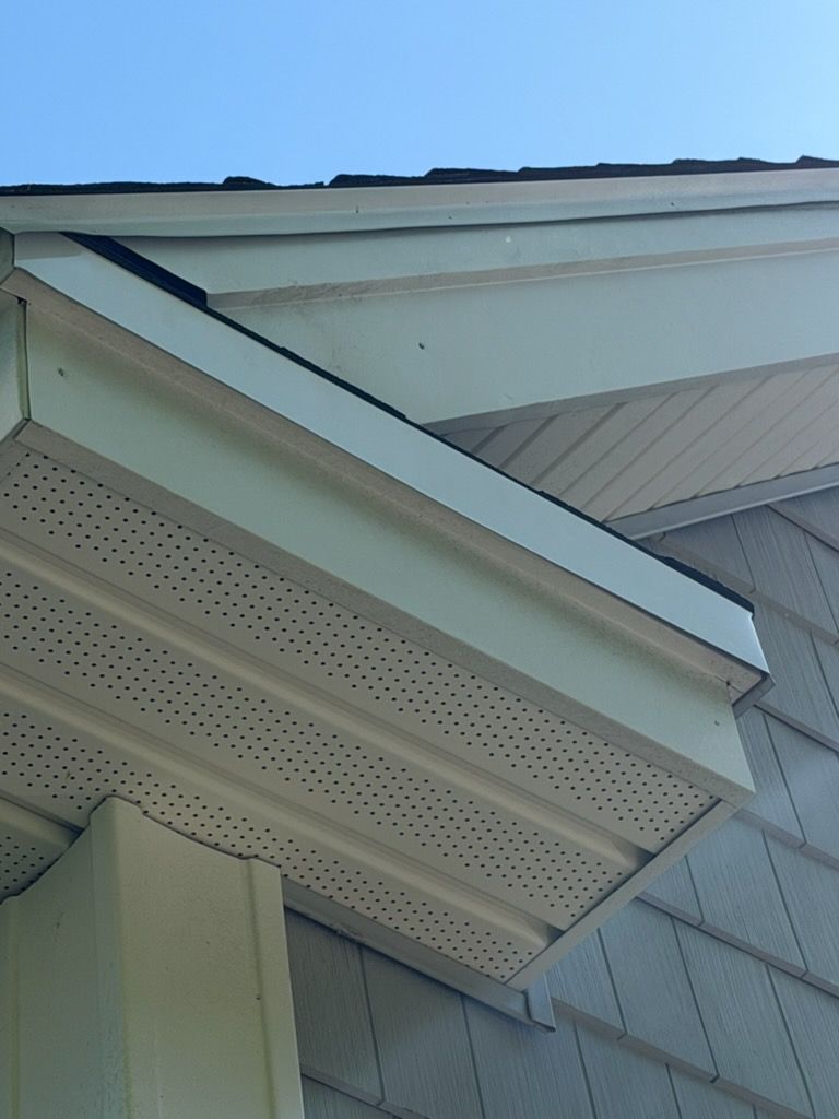 Close-up of a house roof eave with white trim, soffit vents, and gray siding under a blue sky
