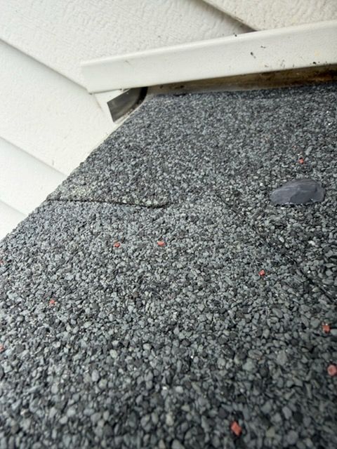 Close-up of damaged roof shingles meeting a white wall, with a crack and a small missing section.