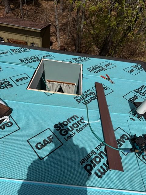 Blue roof with a square skylight opening, roofing underlayment, and a loose board near trees.