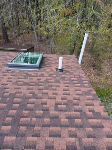 Shingled rooftop with a skylight, vent pipe, and wooded trees in the background