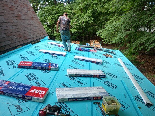 Worker installing blue roofing underlayment on a sloped roof beside trees