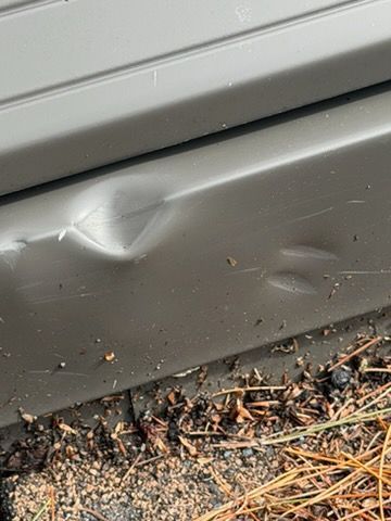 Gray metal panel with a dent near the bottom edge, above dirt and pine needles.